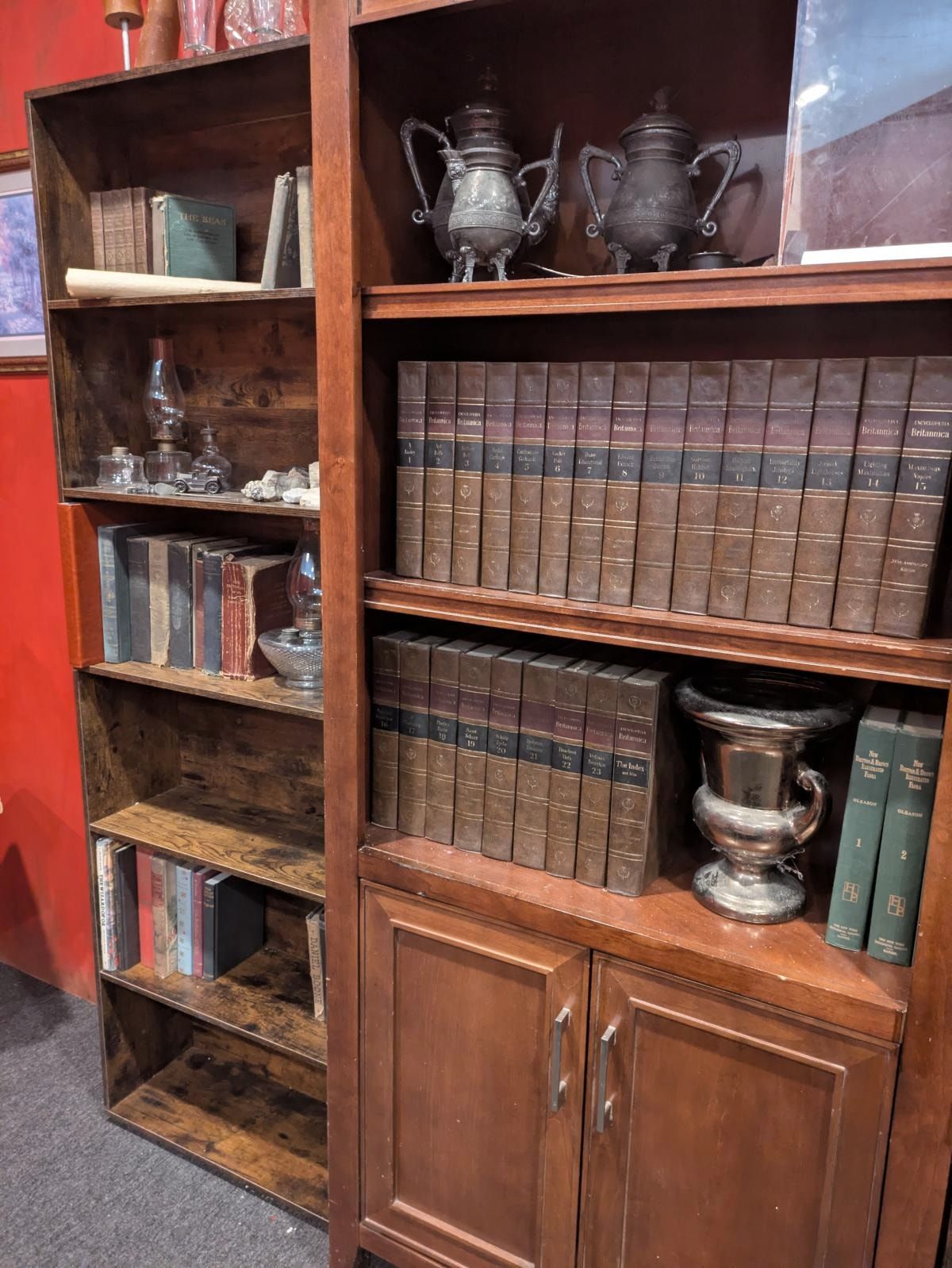 A corner of the parlor: two wooden bookcases against a red wall, holding a full set of Encyclopedia Britannica, tarnished silver urns, oil lamps and rows of worn hardbacks.