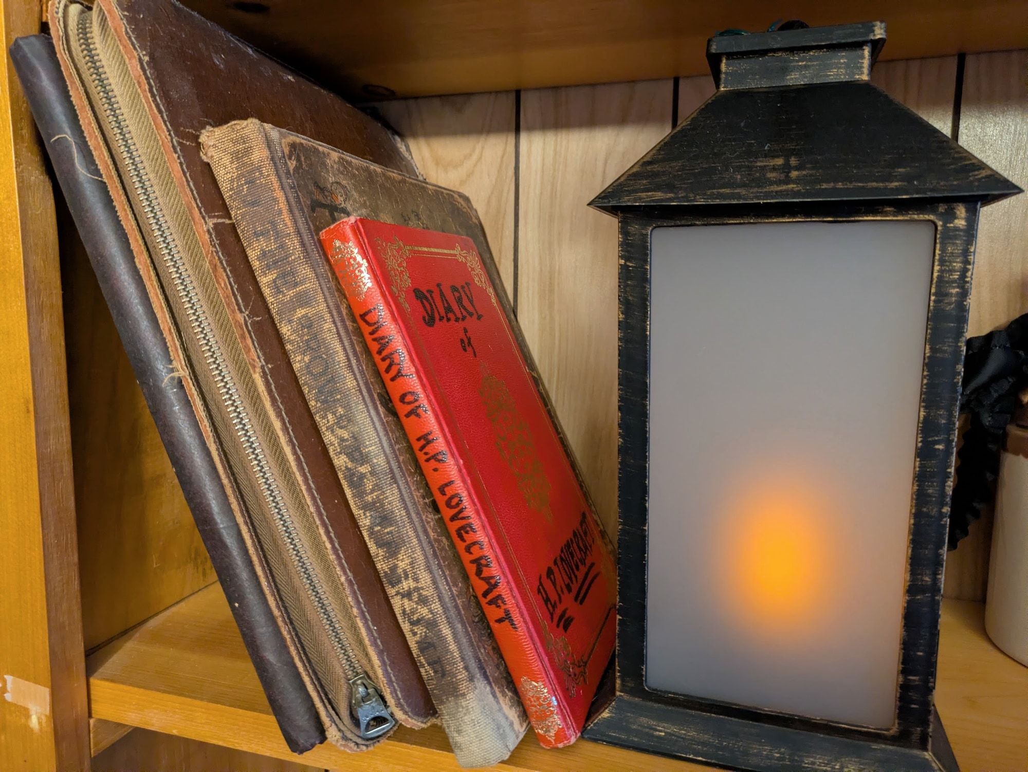 A shelf in the parlor holding worn cloth-bound books and a red gilt-lettered volume titled Diary of H.P. Lovecraft, beside a lit black metal lantern.