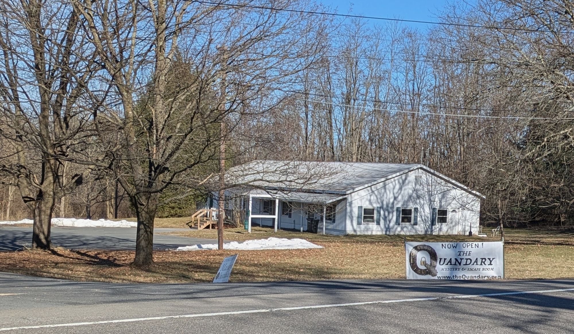 The Quandary from Trumansburg Road: a low white building set back behind bare trees, with a white Now Open banner for The Quandary staked by the roadside.