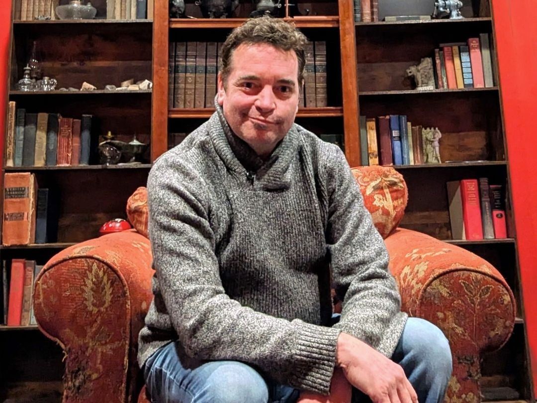 The parlor set: deep red walls, a wall of bookcases holding antique books, glass bottles and oil lamps, a brass telescope on a tripod, and an oil painting of a rocky New England harbour. Daniel Trippett sits in an antique armchair in the middle of it.