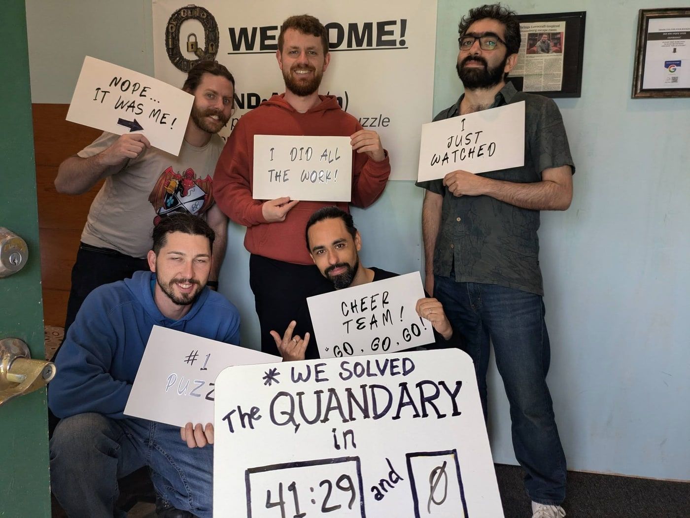 Five men in the lobby, one punching the air, holding hand-lettered boards. The largest reads: We solved The Quandary in 41:29 and 0 hints.