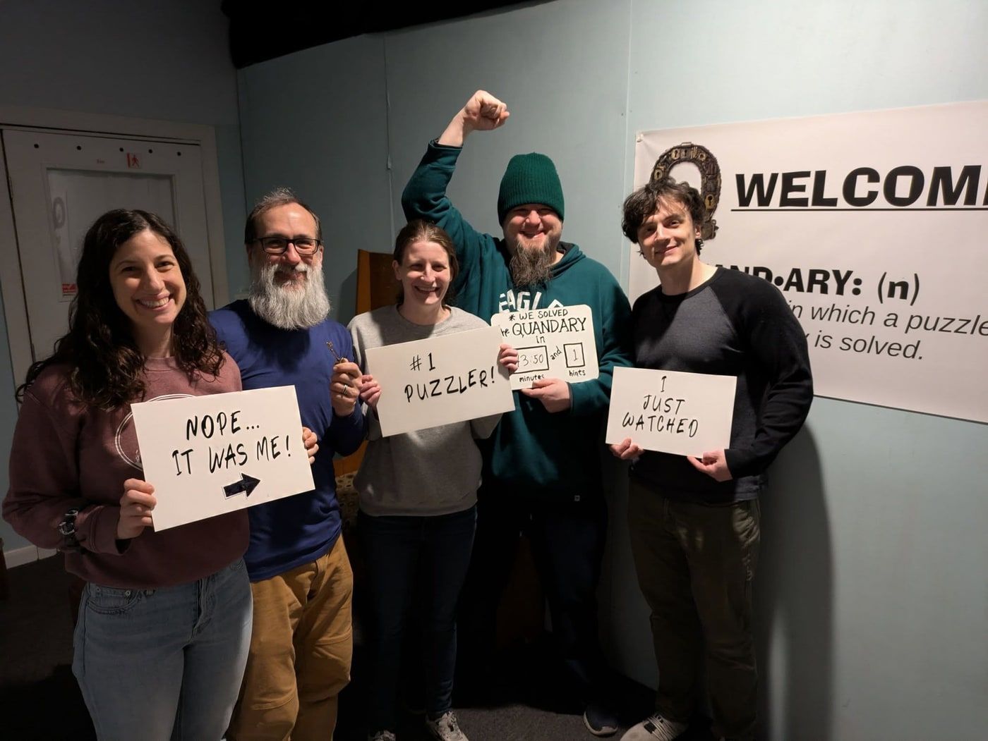 Five players in front of the Welcome banner, one raising a fist, with signs reading #1 Puzzler, Nope… it was me, and I just watched.