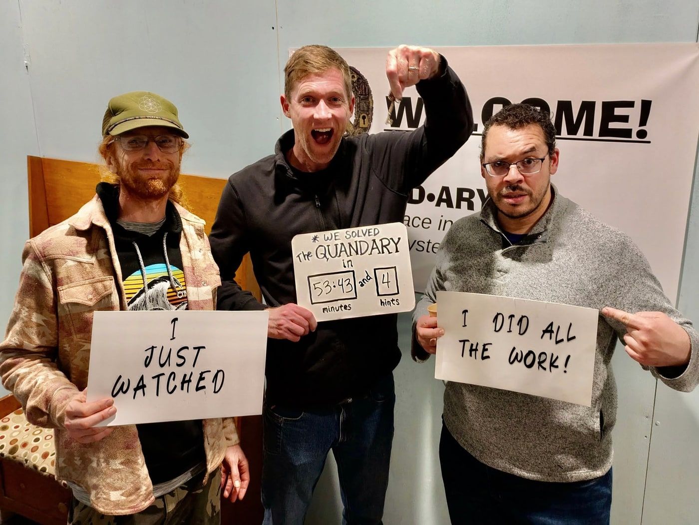 Three men grinning at the camera, one cheering, holding boards reading I just watched and I did all the work, beside their 53:43 and 4 hints scoreboard.