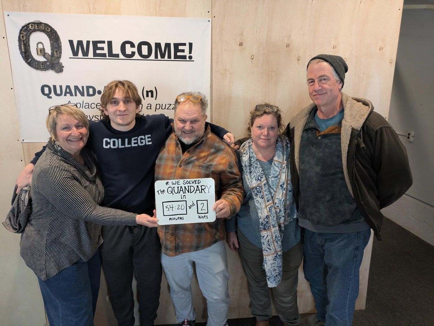 A family of five with arms around each other in front of the Welcome banner, holding a board reading We solved The Quandary in 54:20 and 2 hints.
