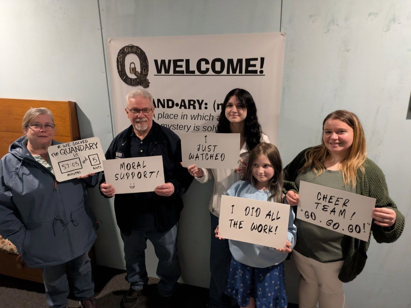 Three generations of one family, including a young child holding a sign reading I did all the work, beside a board showing 57:05 and 5 hints.